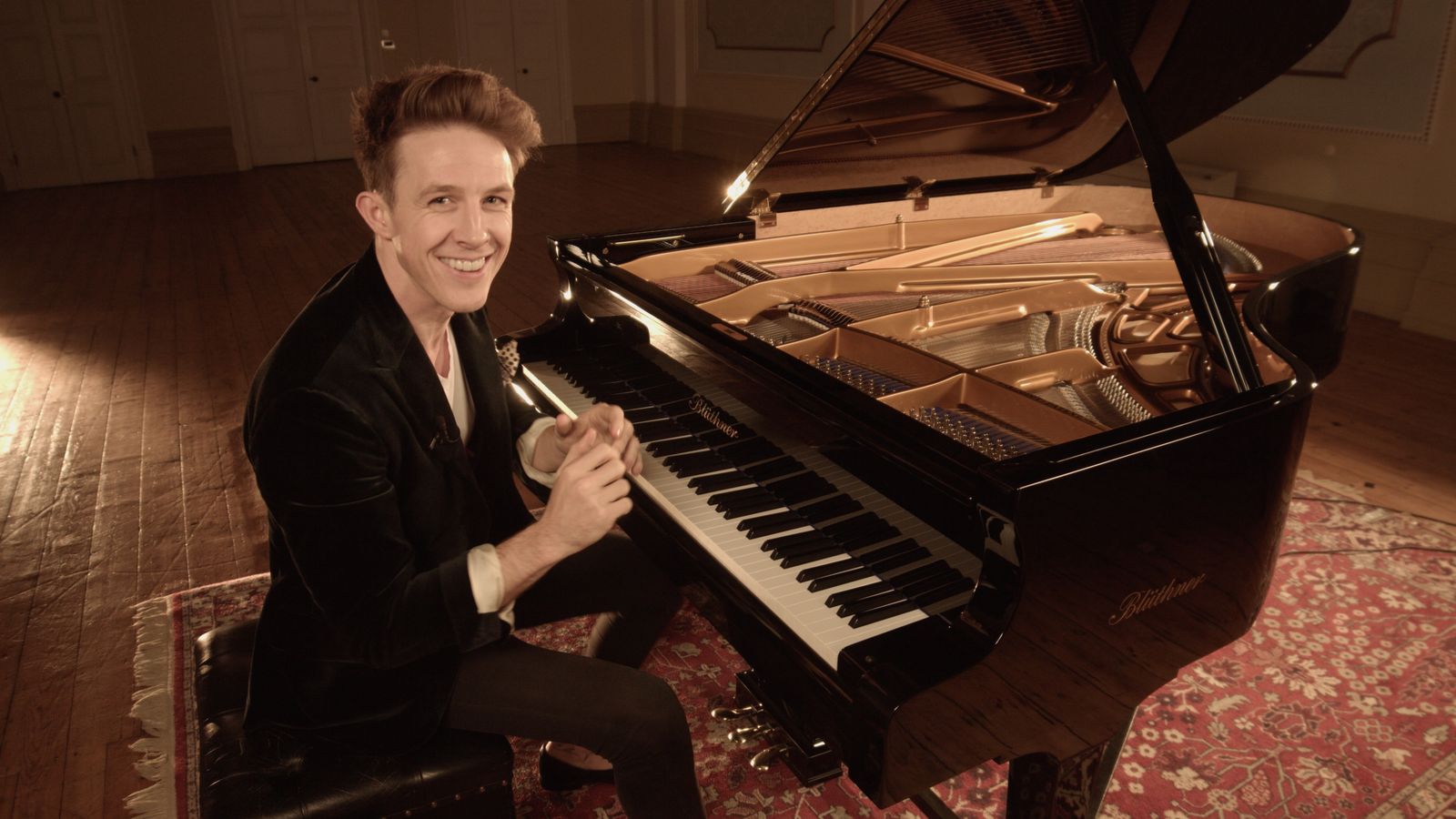 Stephen Ridley seated at an open grand piano in a warm recital hall, smiling at the camera